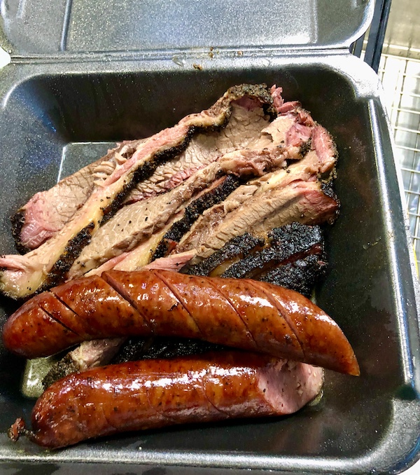 A black take out container with slices of beef and two sausages in Sonora Texas