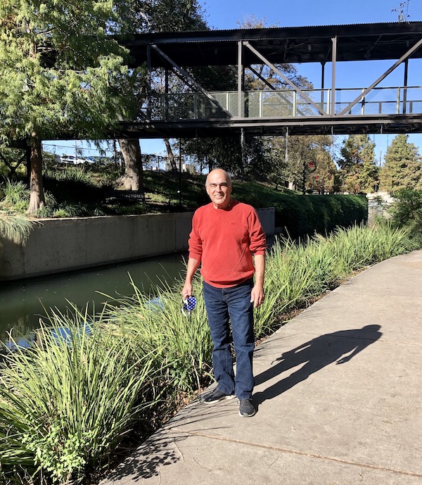 A man in jeans and a rust colored sweater stand by a river in The Pearl in San Antonio