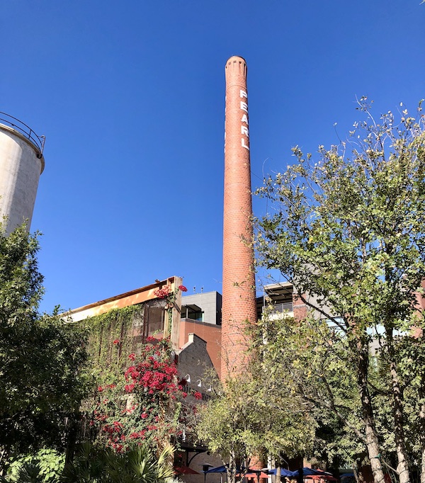 Trees, old buildings and a tall red brick smoke stack with the word Pearl in white on it at The Pearl in San Antonio