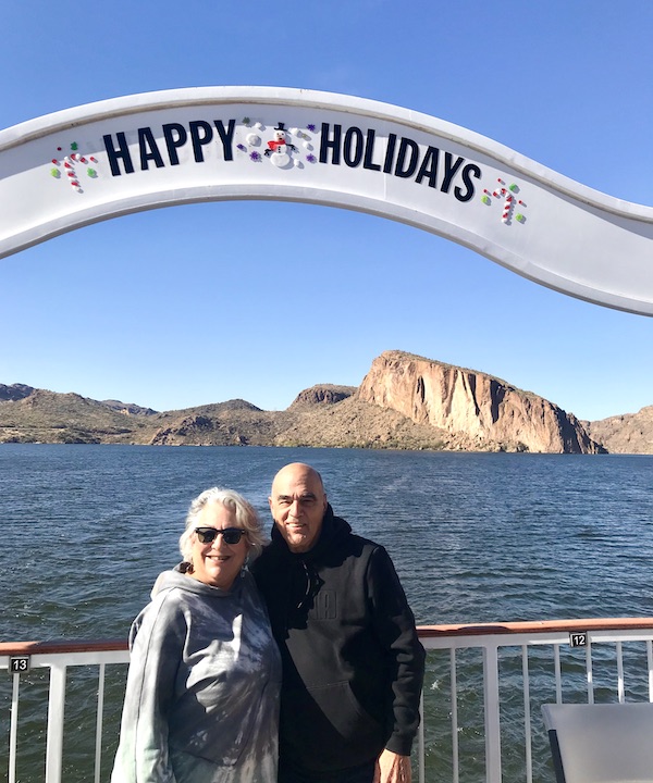 A man and a woman on a steamboat under a white banner that says Happy Holidays. Mountains, lake and blue sky in the background along the Apache Trail Arizona