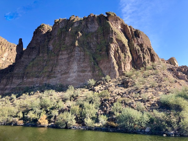 A large rock pops out of the water with green bushes at the base along the Apache Trail Arizona