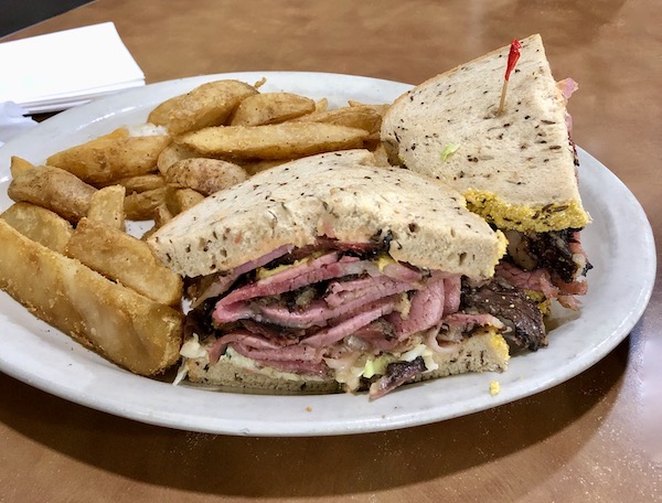 A white plate topped with a red meat sandwich on seeded bread with a side of wide french fries at Chompie's Deli.
