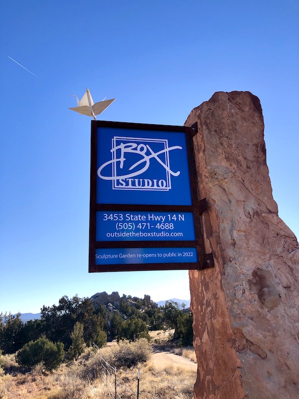 A large tree trunk supports a blue sign for the Box Gallery on our drive from Santa Fe to Albuquerque New Mexico