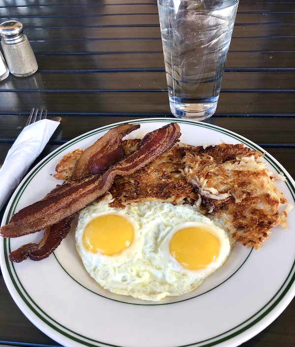 A white plate with a black rim topped with 2 sunny side up eggs, bacon and hash brown potatoes at the Plaza Café in Santa Fe
