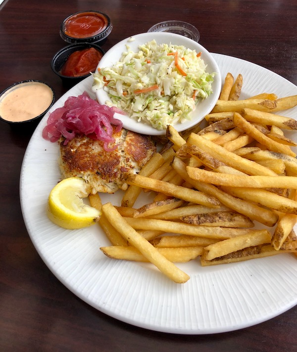 A round white plate topped with a crab cake, french fries, pickled red onion and coleslaw en route Coast to Coast We Did It!