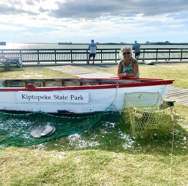 A woman standing behind a white row boat with lake in the background en route Coast to Coast We Did It!
