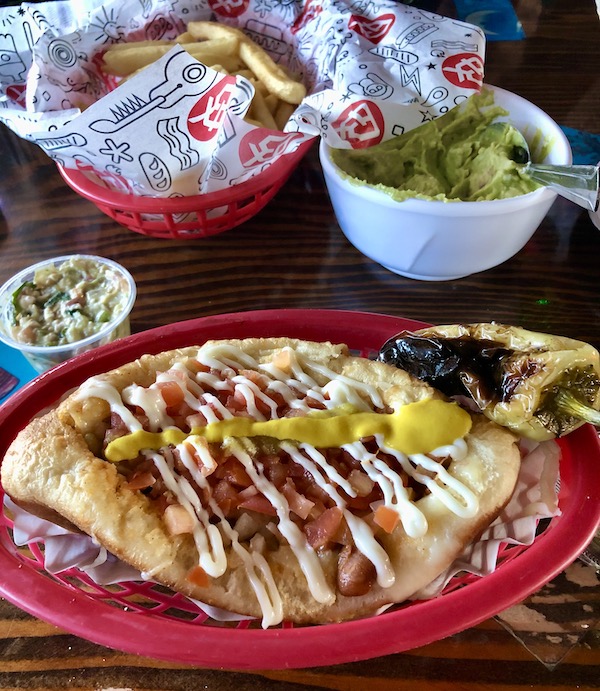 A red platter topped wit a hotdog with many condiments, a white bowl of avocado dip and a red basket of tortilla chips eaten en route Coast to Coast We Did It!