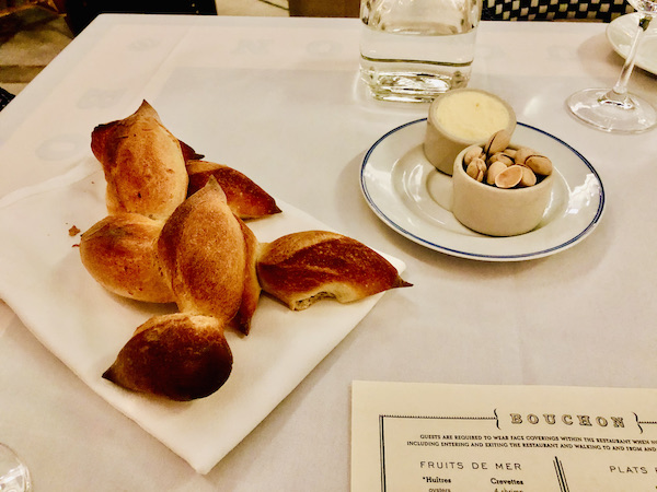 A white square plate topped with football shaped rolls and a small round plate holding a ramekins of butter and pistachios at Thomas Keller's Bouchon Las Vegas