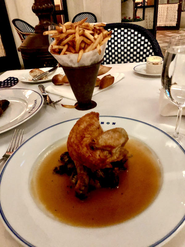 A cream colored plate topped with roasted chicken, Brussels sprouts an jus with a cone of fries in the background at Thomas Keller's Bouchon Las Vegas