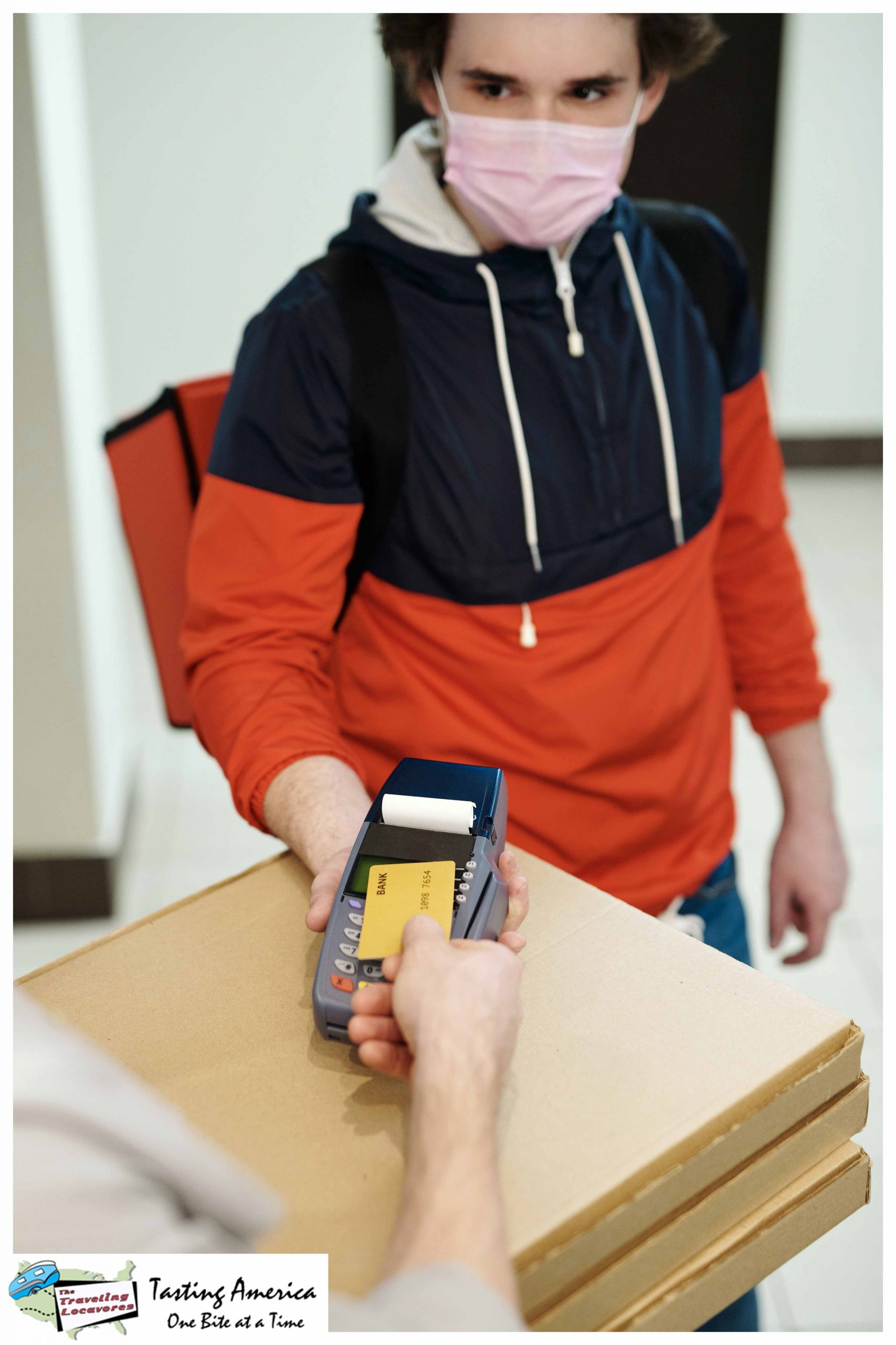 A man in a blue and red hoodie with a pink mask holding a payment terminal is an example of the 5 changes in restaurants caused by the pandemic that are here to stay