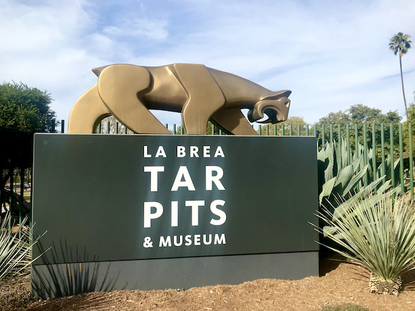 A black marble wall with white lettering welcoming you to the La Brea Tar Pits, with a gold leopard sculpture on top seen during A Day in LaLa Land