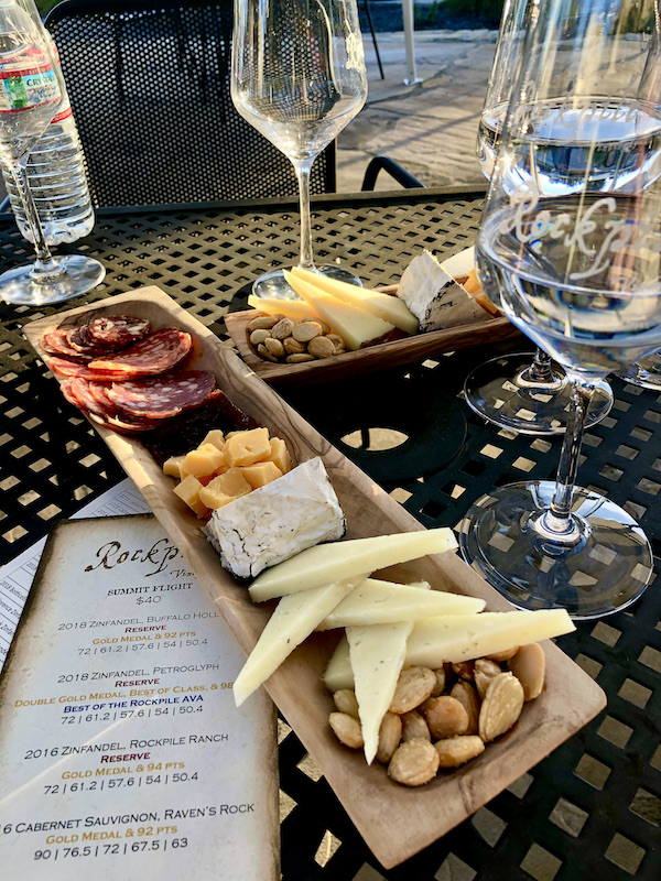 A patio table topped with long, narrow wooden platters and cheeses, salamis, nuts and fruit along with wine glasses enjoyed during A Day with Friends in Windsor California