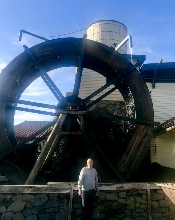A man in a grey sweatshirt standing by a huge waterwheel and white building at Casa de Fruta Hollister California