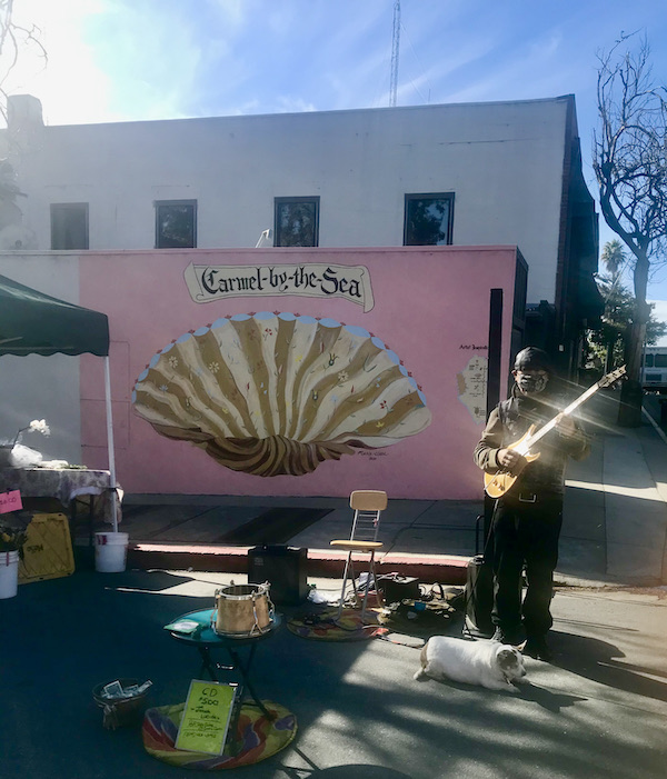A man with a protective mask playing guitar in front of a pink wall with a large scallop shell painted on it. Seen near the Scenic 17-mile Drive Monterey California