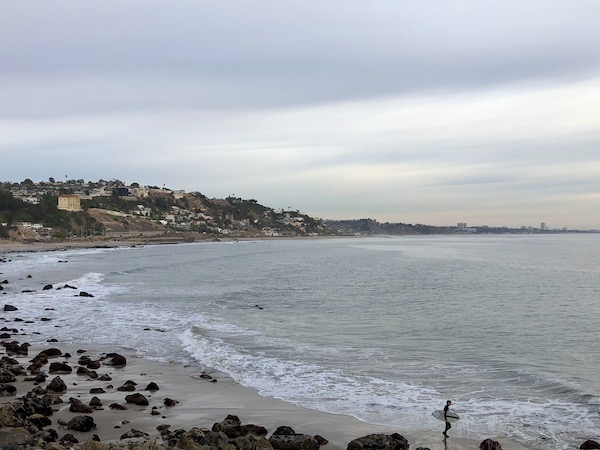 A surfer carrying a surf board, ocean and rocks along the Southern California Coast