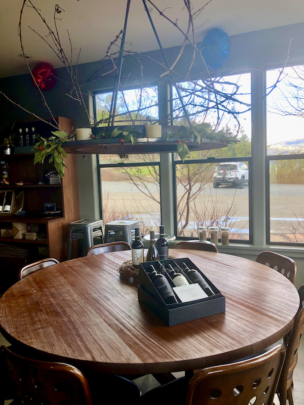 A round wood table by a multi-paned window. Then table is topped with a box showing 3 bottles of wine seen on our tour Behind the Scenes at Stuhlmuller Vineyards