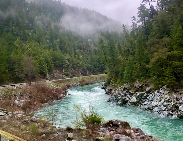Rocks and a green river with lots of trees, mountain behind white cloud on our way to San Francisco California