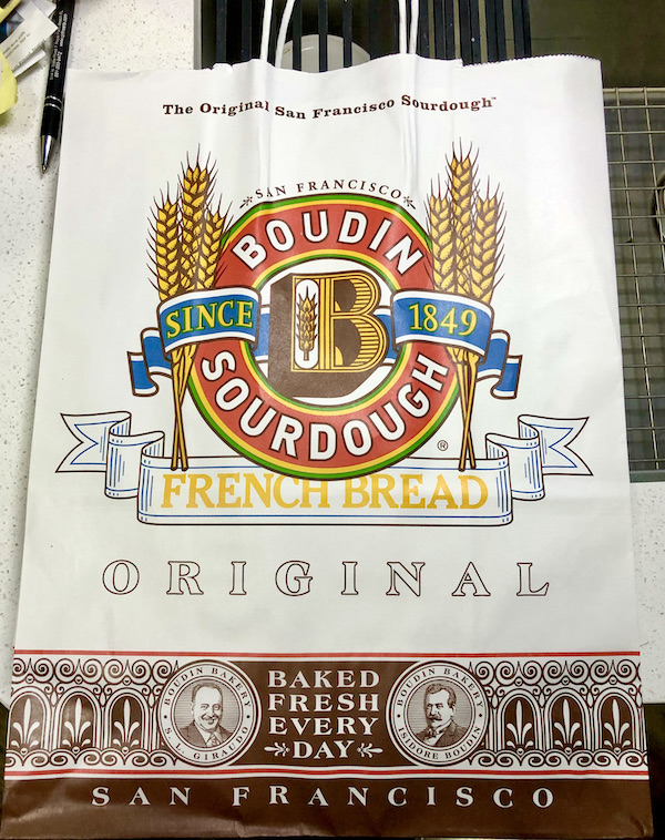 A white shopping bag with the Boudin Bakery logo in god and red from San Francisco California