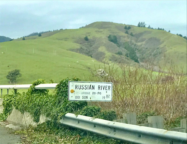 A grassy hill with bushes. In the foreground is a rail and a sign for the Russian River, seen on hour way back from Sonoma Coast State Beach