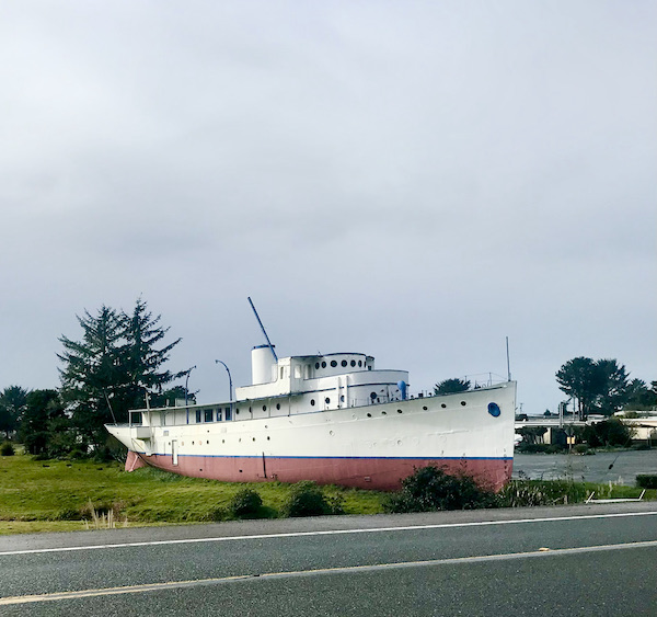 An old wood boat by a street with ocean in background near The Hungry Clam Brookings Oregon