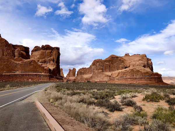 A paved road running passed red rock mountains that look like medieval fortresses at Arches National Park Moab Utah