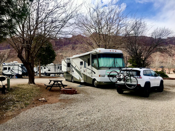 An RV parked between tree and a Jeep with a bike on the back parked in front of the RV near Arches National Park Moab Utah