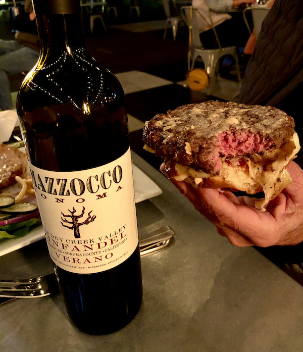 A man's hand holding a burger without a top bun. On the table to the left is a bottle of red wine at Coyote Sonoma in Healdsburg CA