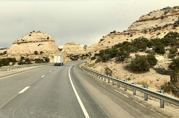 A white box truck on a highway flanked by off white mountains with black stripes and trees on our Scenic Drive to Moab Utah