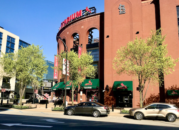 A red brick baseball stadium with a rounded left side. Trees and cars are in front of the building.