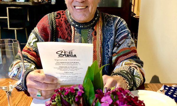 At Stella Modern Italian Cuisine OKC a man in a multicolored sweater is seated at a wood topped table, reading a menu.