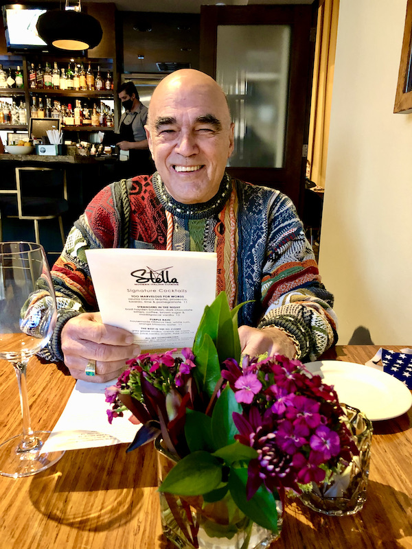 At Stella Modern Italian Cuisine OKC a man in a multicolored sweater is seated at a wood topped table, reading a menu.