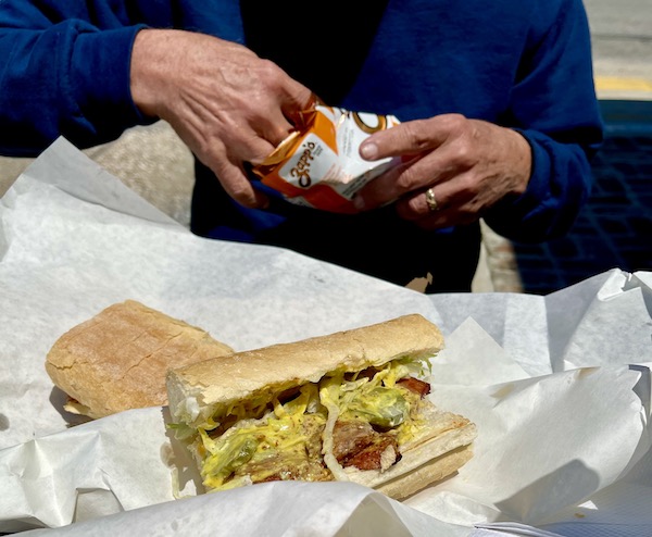 A man taking chips from a bag. In front of him is white deli paper topped with a sausage Po' Boy eaten while Back in NOLA for a Month