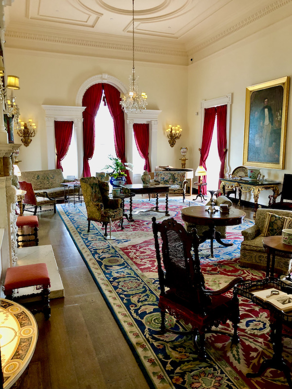 A huge room with pale yellow walls, arched windows with red drapes and a Persian rug. Seating areas with chairs and tables are all in this Drawing Room.