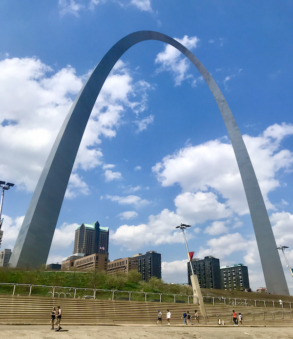 A tall metal arch with buildings and lots of steps. Blue sky with white clouds in background.