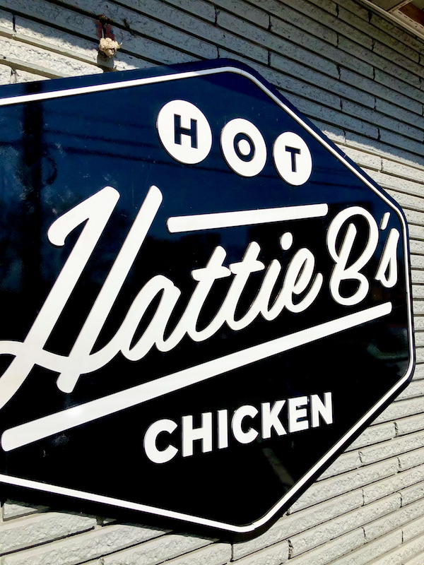 A large black and white sign for Hattie B's Nashville Hot Chicken on a white siding exterior wall.
