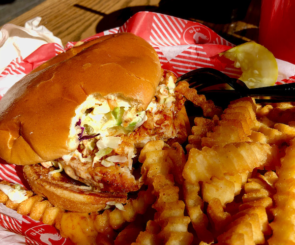 A fried chicken sandwich with coleslaw and fries. A bite has been eaten from this Hattie B's Nashville Hot Chicken sandwich.