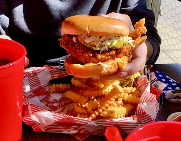 A man's hand holding his Hattie B's Nashville Hot Chicken sandwich. above crinkle fries in a red and white checkered lined basket.