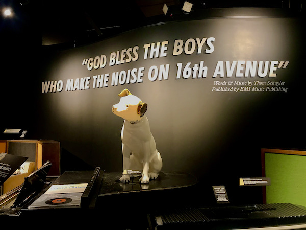 A small white dog with black ears and head cocked to one side sitting against a black wall at the Musicians Hall of Fame Nashville