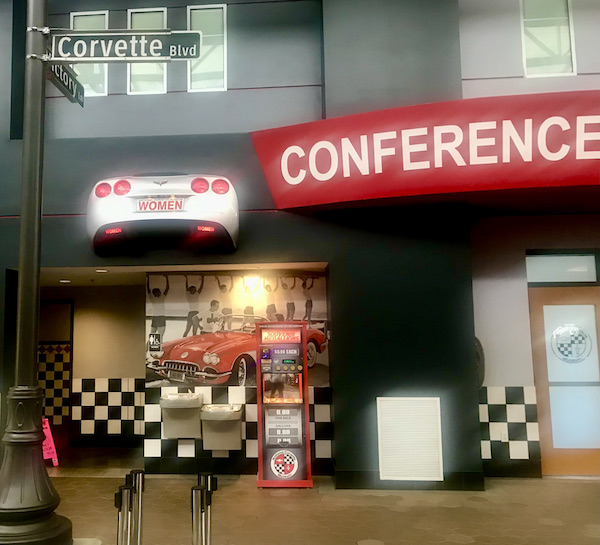 The rear of a silver Corvette with a license plate that says Women at the checkered entrance to a bathroom at the National Corvette Museum