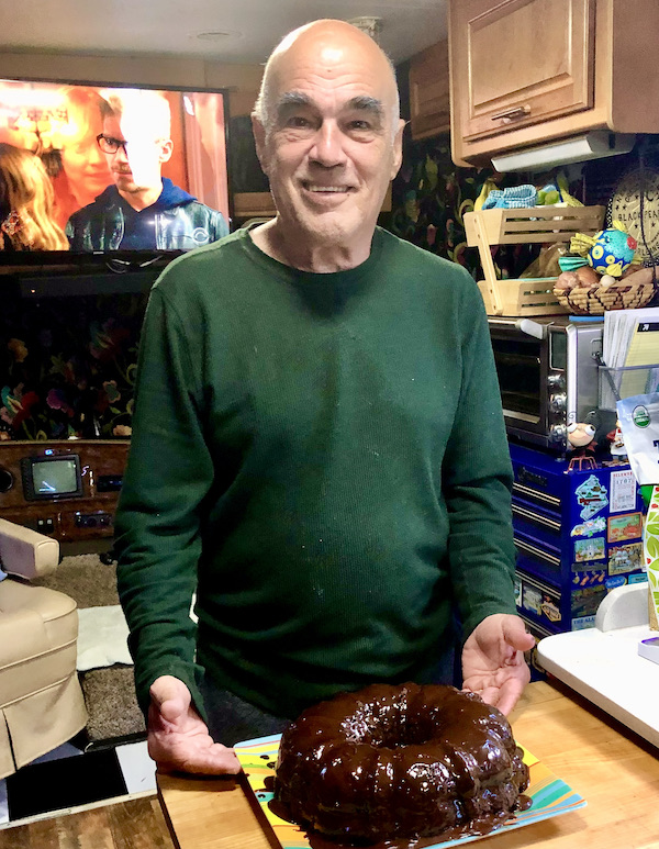 A man in a dark green sweater presenting a chocolate bundt cake on a striped plate.