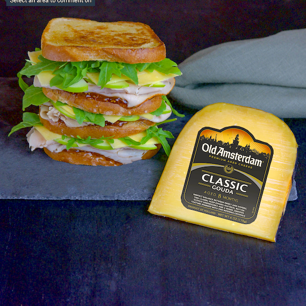 A tall stacked sandwich with cheese sitting on a blue cloth along with a wrapped wedge of Old Amsterdam Cheeses.