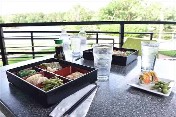 A black bento box filled with Asian food, glasses filled with water and other dishes on a table overlooking Japanese Gardens