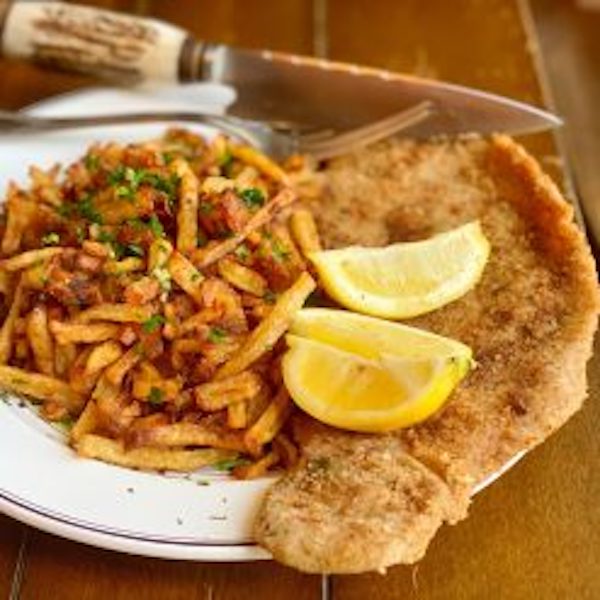 A white plate topped with a piece of breaded beef, two lemon wedges and fries from Sobremesa - A Memoir of Food and Love