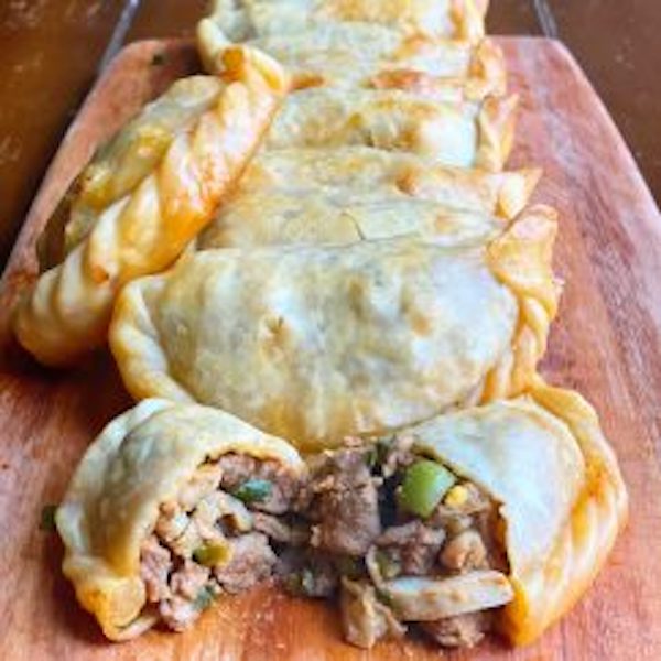 A wood cutting board topped with several empanadas with the front one cut open from Sobremesa - A Memoir of Food and Love