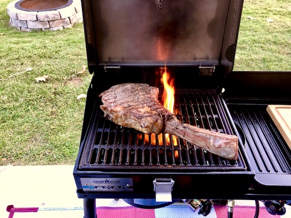 A large Tomahawk Steak on the Grill with a small flame shooting out.