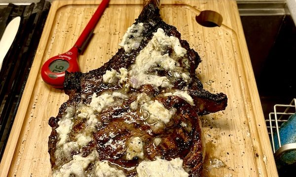 A cooked Tomahawk Steak on the Grill with butter melting on it and a red meat thermometer sitting on a cutting board.