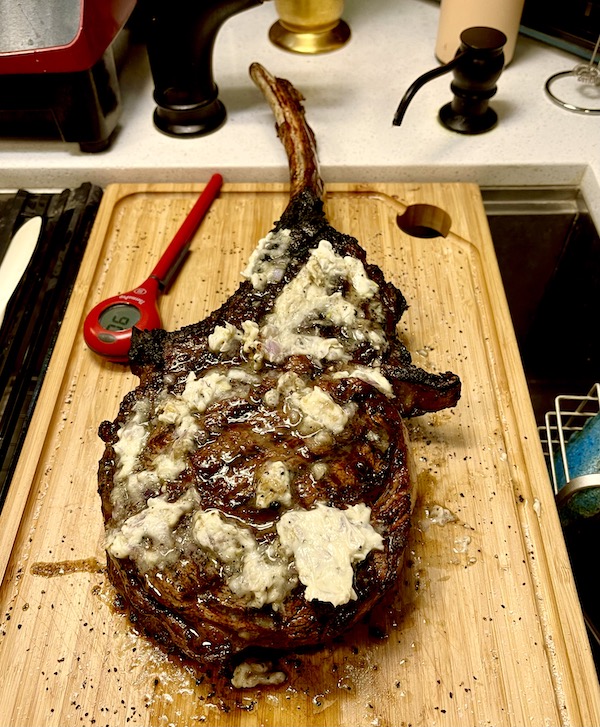 A cooked Tomahawk Steak on the Grill with butter melting on it and a red meat thermometer sitting on a cutting board.
