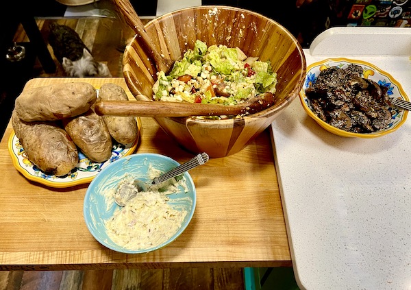 A wood bowl of salad, baked potatoes, mushrooms and butter topping for Tomahawk Steak on the Grill