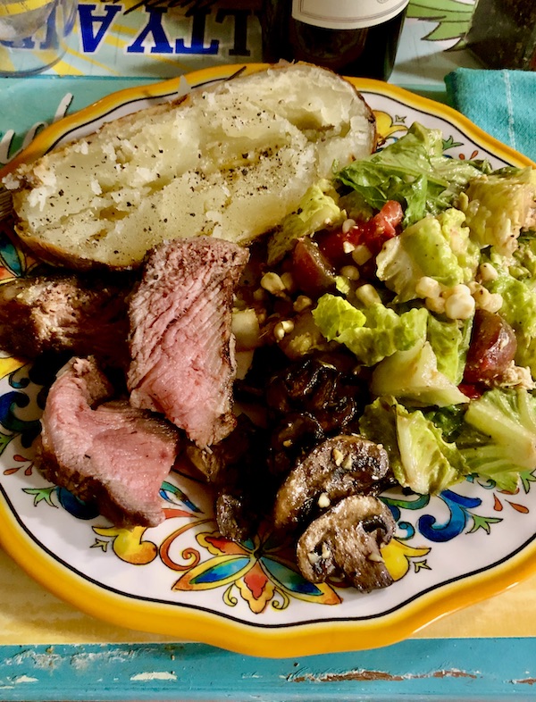 A plate with a yellow rim topped with baked potato half, steak, mushrooms and salad. A perfect Tomahawk Steak on the Grill dinner.