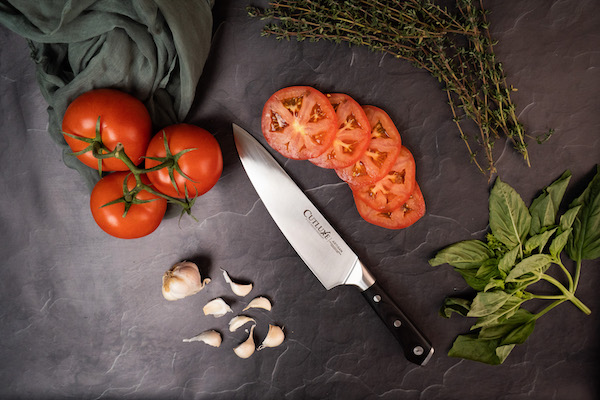 A chef knife on a dark wood surface with sliced and whole tomatoes and garlic cloves from 15 Best Gifts for Foodies and RVers